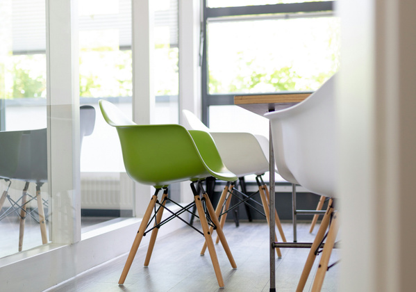 KI generiert: Moderne, bunte Designerstühle in einem hellen Raum mit großen Fenstern.