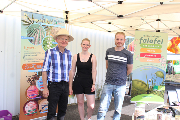 KI generiert: Drei Personen vor Falafel-Werbung auf einem Marktstand. Text: "Sachsenhäuser falafel", "Gut-Fränkisch", "Arabica".