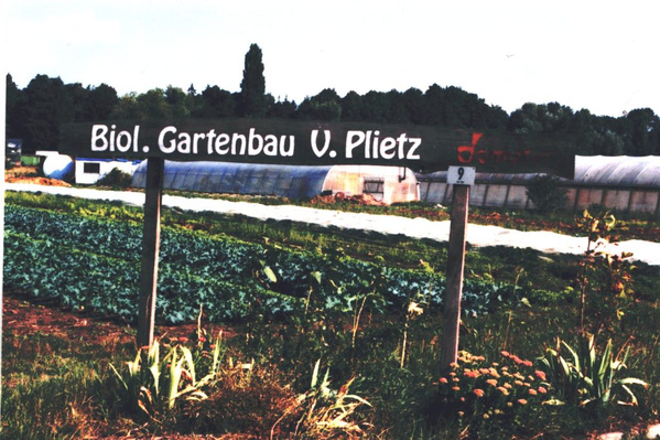 KI generiert: Ein Schild mit "Biol. Gartenbau V. Plietz" vor einem Feld mit Gewächshäusern und Pflanzen.
