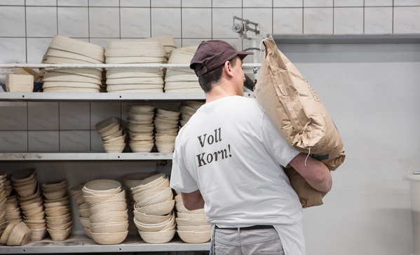 KI generiert: Ein Bäcker trägt einen Mehlsack. Text auf dem T-Shirt: "Voll Korn!"