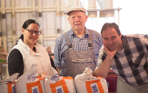 KI generiert: Drei Personen stehen hinter Säcken mit Aufschrift "WE BA". Sie lächeln in die Kamera in industrieller Umgebung.