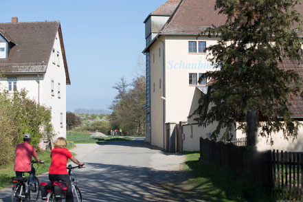 KI generiert: Zwei Personen radeln auf einer Landstraße zwischen Häusern. Im Hintergrund steht "Schaub..." auf einem Gebäude.