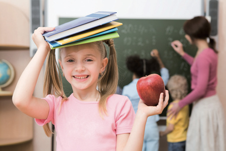 KI generiert: Im Vordergrund ist ein lachendes Mädchen, das Bücher auf dem Kopf balanciert und einen Apfel hält. Im Hintergrund unterrichtet eine Frau zwei weitere Kinder an einer Tafel.