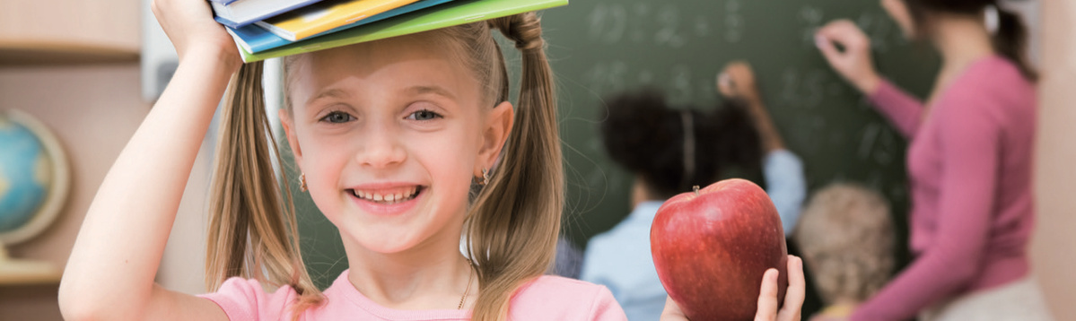 KI generiert: Im Vordergrund ist ein lachendes Mädchen, das Bücher auf dem Kopf balanciert und einen Apfel hält. Im Hintergrund unterrichtet eine Frau zwei weitere Kinder an einer Tafel.