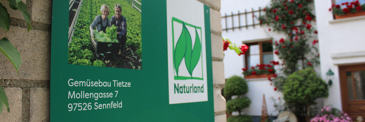 KI generiert: Schild von "Gemüsebau Tietze" mit Naturland-Logo und Foto von zwei Menschen in einem Gemüsegarten.