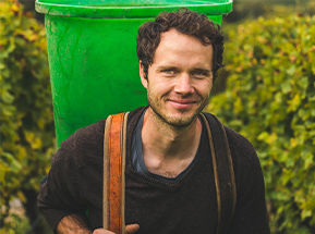 KI generiert: Ein Mann trägt einen großen, grünen Behälter auf dem Rücken, umgeben von Pflanzen.