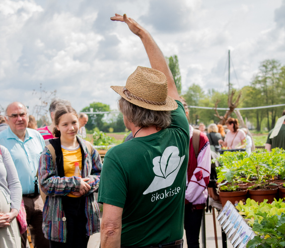 KI generiert: Eine Person mit Hut erklärt einer Gruppe etwas auf einem Gelände. Text: "ökokiste".
