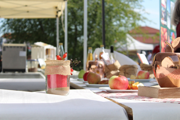 KI generiert: Ein Tisch mit dekorativen Gläsern, Äpfeln und Essensproben auf einem Freiluft-Markt.
