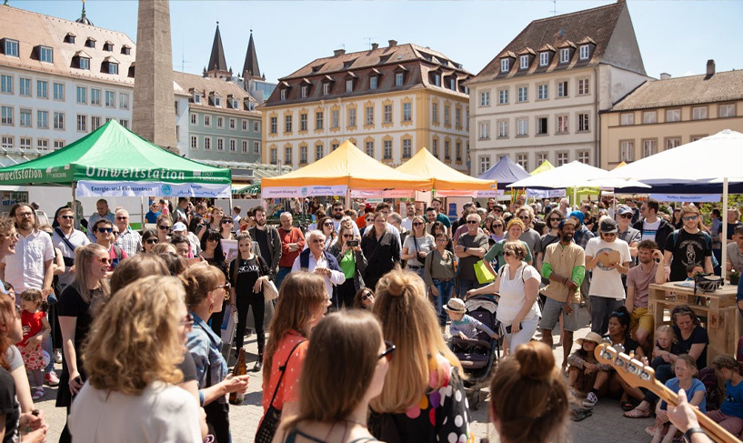 KI generiert: Menschenmenge auf belebtem Platz mit bunten Marktständen und einem Zelt mit "Umweltstation" im Hintergrund.