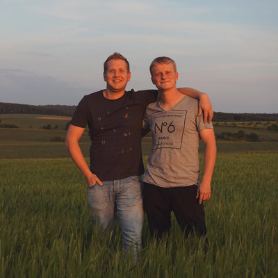 KI generiert: Zwei Männer stehen lächelnd auf einem Feld, im Hintergrund eine Landschaft bei Sonnenuntergang.